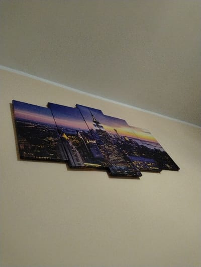An interior scene in Oreshene, Bulgaria, presents a multi-panel canvas print mounted on a light beige wall. The print, composed of several rectangular sections of varying dimensions, displays an urban cityscape at twilight or dawn. The depicted scene features numerous illuminated buildings and skyscrapers, including a prominent rendering of the Empire State Building, set against a sky that transitions from dark blue at its zenith to orange and yellow near the horizon. The panels are horizontally aligned and staggered, with a smaller, lower panel positioned centrally to the right. Above the artwork, the light beige wall extends upward to meet a similarly colored ceiling, separated by a distinct white trim line. No individuals or other objects are visible within the frame.
