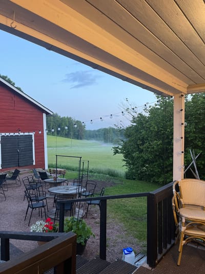 The image depicts an outdoor seating area next to a red barn on a clear, early morning. The setting appears to be a rural or semi-rural area in Vantaa, Finland, likely an establishment with outdoor dining or a gathering space.

In the foreground, a dark wooden porch railing overlooks a gravel patio with several black metal chairs and round wooden tables. Two potted plants, one with red flowers and another with green foliage, are placed on the patio. A blue-capped plastic jug sits on the porch floor near the railing.

The red barn, a prominent structure on the left, features large, dark double doors and white trim. String lights with exposed bulbs are draped across the porch and along the side of the barn.

Beyond the seating area, a vast, misty green field stretches towards a treeline in the distance. The mist creates a soft, atmospheric effect, obscuring the details of the landscape. Several streetlights are visible along a road winding through the field. The sky is a pale blue, transitioning from dawn, with a few wispy clouds. The overall impression is one of tranquility and a peaceful start to the day.