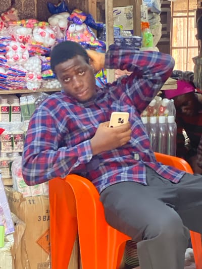 A man in a plaid shirt and grey pants leans back in an orange plastic chair, holding a phone. He appears to be in a shop or market, surrounded by various items for sale including packages of marshmallows, bottled drinks, and boxes labelled "FIREWORKS." Another person is visible in the background, also in a plaid shirt. The setting suggests a busy, informal commercial environment.