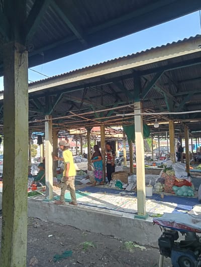 The image depicts an outdoor market scene in Ujung Tanjung, Indonesia. The market is covered by a corrugated metal roof supported by wooden and concrete pillars, with some parts showing wear and tear. Sunlight filters through the structure, illuminating a bright blue sky.
In the foreground, a man wearing a yellow t-shirt and beige pants walks away from the camera, his back to the viewer. He is on a concrete walkway overlooking a muddy area with some scattered green leaves.
Further into the market, several people are visible. One woman in a blue and pink patterned dress stands holding a basket, interacting with another person whose back is to the camera. To the left, another woman in a green top is seated behind a stall displaying various vegetables, including green beans and potatoes.
The market stalls are arranged with goods laid out on blue and white striped cloths. Piles of vegetables, such as carrots, cabbages, and onions, are prominently displayed. Woven baskets and plastic bags are used to hold and transport the produce.
In the background, other market stalls, vehicles, and buildings can be seen, suggesting a bustling commercial area. The overall impression is of a typical Indonesian market, with vibrant colors and active commerce under a shaded canopy. A blue generator or engine is visible in the lower right corner of the image.