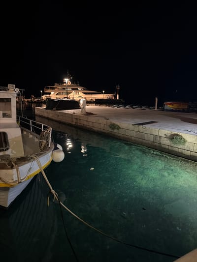 The image depicts a night scene in a harbor, likely in Općina Brela, Croatia. The primary subjects are boats docked along a stone quay. The water is clear and greenish, with reflections of lights visible on its surface. The quay is illuminated by artificial lights, casting long shadows.
On the left side of the image, a large, multi-story vessel is moored. It appears to be a ferry or a cruise ship, with multiple decks and lit windows. Moored closer to the foreground is a smaller boat, possibly a fishing boat or a pleasure craft. It has a white hull with yellow accents and is equipped with safety equipment like lifebuoys and ropes. One of the lifebuoys is prominently white and spherical.
The scene is dark, indicating it's nighttime. The weather appears calm, as the water is still and shows clear reflections. The presence of a harbor, the style of the quay, and the water suggest a coastal location. There is a hint of graffiti on a signpost attached to the quay wall. No people are visible in the frame. The overall mood is tranquil and still, typical of a harbor at night.