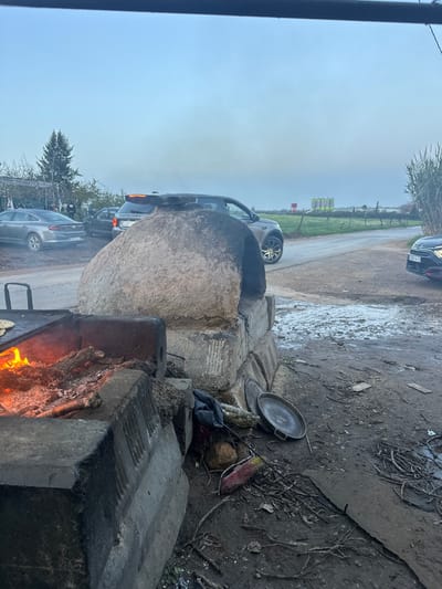 Outdoor scene with an earthen oven and a nearby barbecue. The oven is large and rounded, situated near a row of parked cars on a road. A barbecue is burning with fire visible in the lower foreground. Corn is positioned near the oven. The surrounding environment appears to be rural or semi-rural, with trees and sky visible in the distance. The overall setting suggests an outdoor cooking or gathering space.