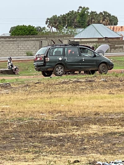 An outdoor, medium shot captures a dark green minivan with its hood up, seemingly undergoing maintenance, in a semi-urban or rural environment in Wai A, Nigeria. The minivan is parked on a dirt and grass area, with its rear slightly damaged. A person in a patterned headscarf and dress is seated on a concrete block to the left of the vehicle, facing away from the camera. In the background, a stone wall and buildings with corrugated metal roofs are visible, along with a cluster of palm trees. The ground in the foreground is dry and covered with sparse vegetation. The sky is overcast.