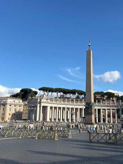 The image depicts St. Peter's Square in Città del Vaticano, Vatican City, under a clear blue sky with scattered white cumulus clouds. The foreground features a large cobblestone plaza partially sectioned off by light-colored, crisscross-patterned barriers, through which numerous pedestrians are walking. Rows of dark, cylindrical bollards are visible behind these barriers.
Dominating the central midground is a tall, unadorned Egyptian obelisk, topped with a spherical element and a cross. The obelisk casts a long shadow towards the right. On either side, the semicircular colonnade of St. Peter's Square is prominent, composed of multiple rows of classical white columns supporting an entablature lined with numerous statues of saints. Behind the left side of the colonnade, multi-story buildings with light-colored facades and rectangular windows are visible. Elevated beyond the colonnades, the horizon is punctuated by dense clusters of flat-topped umbrella pine trees. Sunlight illuminates the scene from the left, casting shadows from the architectural elements and people onto the square's pavement. People are moving throughout the square, primarily walking, and appear to be visitors or tourists.