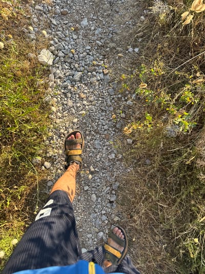 In Brajići, Montenegro, a person's feet, clad in dark shorts and sandals, are positioned on a gravel path. To the left, green grass and dry vegetation border the path, while to the right, a dense growth of dry grass and green foliage creates a natural boundary. The sun casts shadows, indicating it is likely daytime. The scene captures a moment of walking in a natural, outdoor environment.
