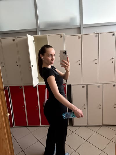 The image shows a woman taking a selfie in a locker room. She is wearing a black athletic outfit and has her hair in a ponytail. She is holding a smartphone in her right hand and a water bottle in her left hand. The locker room appears to be clean and well-lit, with white and red lockers. There are numbers on some of the lockers, including "2", "3", and "4". The floor is tiled with a diamond pattern. The woman is standing in front of a row of lockers, with one locker door open. The scene does not provide any specific information that would indicate the location in the city of Pushkino, Russia.