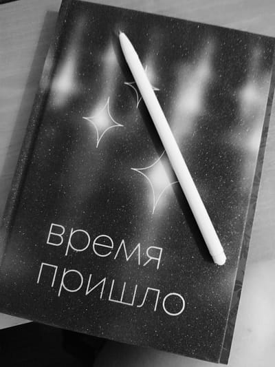The image displays a close-up, overhead view of a black notebook with a starry design. A white stylus rests diagonally across the notebook's cover. The notebook's title, written in white Cyrillic letters, appears to read "время пришло" which translates to "the time has come." The background consists of a light-colored wooden surface. There is no information to suggest this image is located in Skadovsk, Ukraine.