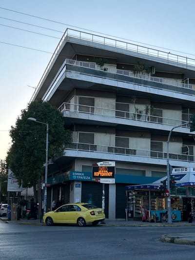 An outdoor, eye-level shot captures a multi-story building and a street scene in Koridallos, Greece. The dominant subject is a modern-style building with several balconies that feature white railings and some potted plants. The building's facade is characterized by alternating light and dark panels. The sky above is clear, suggesting daytime, possibly late afternoon or early morning given the light.
On the ground floor of the building, there are storefronts. A prominent digital sign displays Greek text, reading "ΔΗΜΟΣ ΚΟΡΥΔΑΛΛΟΥ" (Municipality of Koridallos), with "www.korydallos.gr" below it. Another sign for "ΕΘΝΙΚΗ ΤΡΑΠΕΖΑ" (National Bank) is visible further down. An awning or sign for "MAGIC BOX" can also be seen.
A bright yellow compact car is parked on the street next to the curb, in front of the ground-level businesses. A large, leafy tree stands in the foreground, partially obscuring the lower part of the building and some street infrastructure, including a streetlight. Utility wires are visible stretched across the sky. The overall scene depicts a typical urban environment in Greece.