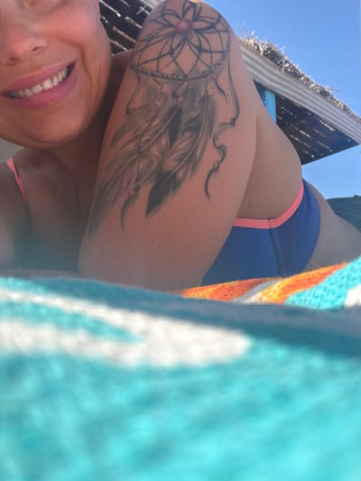A woman is pictured from a low angle while reclining outdoors. She has a fair complexion and is smiling, showcasing her teeth. She is wearing a bikini top and blue bottoms with a pink trim. The woman has a tattoo of a dreamcatcher on her upper arm. A turquoise and white striped towel with orange accents is visible in the foreground. The background includes a partially visible structure with a thatched roof and the clear blue sky, indicating a sunny day. The location is Al Hadaba, Egypt.