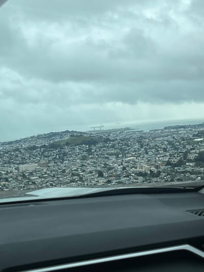 An aerial view shows a dense cityscape under a cloudy sky. Buildings stretch across the landscape toward a body of water, where a bridge-like structure is visible in the distance. The foreground includes a portion of an interior, possibly a car, with dark gray and silver elements.