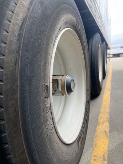 The image shows several large truck tires stacked inside a trailer or cargo area. The tires are black with white rims and have "MICHELIN" and other tire information visible on their sides. One tire is in the foreground, taking up most of the frame, with its tread clearly visible. The tires are arranged in a row, suggesting they are being transported or stored. The background shows the interior of the trailer, with metal surfaces and a yellow line painted on the floor, possibly indicating a parking or loading zone. The lighting suggests it is daytime. There is no visible human presence.