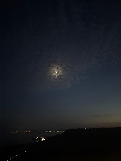 A crescent moon hangs in the night sky, partially obscured by wispy clouds. Below, the dark silhouette of land borders a body of water, where distant lights twinkle. Some of these lights appear to be from boats, while others suggest a settlement or town along the shore. The overall scene is dark and atmospheric, captured at night in the vicinity of Topola, Bulgaria.
