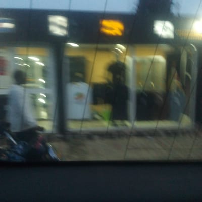 The image, taken from inside a vehicle, depicts a shopfront. A person wearing a light-colored shirt is seated on what appears to be a motorcycle in the foreground. The shop window displays figures, one wearing a white shirt with a design and another in dark clothing. The shop front has lit signs, with one sign displaying what appears to be a logo in yellow. Based on the presence of people, a shop with window displays, and a motorcycle parked outside, the location seems to be a commercial area, possibly a street scene.