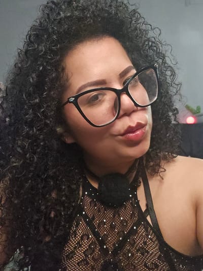 A close-up, frontal, waist-up photograph shows a person with dark, curly hair and glasses. The person is wearing a black, sheer top adorned with rhinestones and a black choker around their neck. The background is a plain, gray wall, with some blurry lights visible on the right side. There is no information in the image to suggest a location in Perímetro Urbano Cúcuta, Colombia.