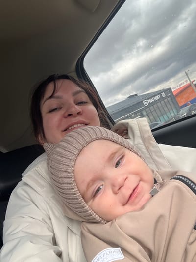A selfie-style photograph captures a woman and a young child inside a vehicle, likely in Protasy, Russia. The image is taken from a low angle, looking upwards through the car's sunroof or window. Both individuals are smiling broadly, looking directly at the camera.
The woman, with dark hair, is seen smiling, revealing her teeth. She wears a light-colored jacket over a dark top. The child, likely a baby or toddler, wears a light beige knitted hat and a matching beige jacket. The child's jacket features a white patch with "ANNA PERIN KIDS" visible on it.
Through the vehicle's sunroof, an overcast, cloudy sky is visible, with raindrops evident on the glass, suggesting wet weather. Beyond the vehicle, reflections of buildings are visible. One prominent reflection shows a building with the Cyrillic text "МАТНИМ 24" which is a reversed reflection of "МАГНИТ 24," indicating a 24-hour Magnit supermarket, a major Russian retail chain. Another building with an orange-red roof is also faintly visible. The scene conveys a cheerful and intimate moment despite the gloomy outdoor conditions.