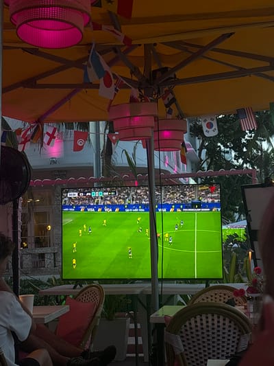 This image captures a lively outdoor scene, likely in Miami Beach, where people are gathered to watch a soccer match on a large screen. The setting appears to be a vibrant open-air venue, possibly a bar or restaurant, adorned with numerous national flags, suggesting a festive atmosphere related to an international sporting event like the FIFA World Cup.
The main subjects are the viewers, partially visible in the foreground, relaxing and focused on the game. We see legs and torsos of people seated on chairs and potentially on the ground. The chairs themselves are of a woven, outdoor style, adding to the casual, tropical ambiance. Hanging decorations, including pink, circular light fixtures and strings of small flags, contribute to the celebratory mood.
The screen prominently displays a soccer game in progress, with green turf and players in yellow and blue uniforms. Text on the screen indicates the match details: "54:04 JPN 0 0 SWE," suggesting the game is in its second half with Japan and Sweden tied. Other visible text includes "FIFA WORLD CUP 2020" and "VISA" advertisements, confirming the event and sponsorship.
The lighting suggests it might be late afternoon or early evening, as the outdoor lighting is illuminated, and the sky is not completely dark. The presence of palm trees in the background hints at a warm-weather location, consistent with Miami Beach. The overall impression is one of shared enjoyment and communal engagement with the sporting event, set against a visually rich and festive backdrop.