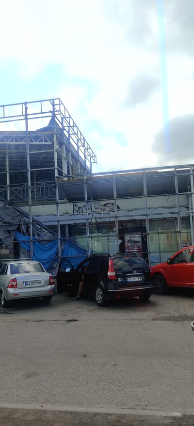 The image depicts a street scene in Valky, Ukraine, with a partially constructed or damaged building in the background. The building appears to be a commercial structure with large, reflective windows, some of which are covered with blue tarps. A metal frame structure is visible extending above the main building. In the foreground, three cars are parked on an asphalt road. A silver sedan is on the left, a black hatchback is in the center with its driver's door open and a person partially visible inside, and a red SUV is on the right. The road shows signs of wear, with some cracks and potholes. The sky is cloudy with a bright lens flare visible towards the upper right.