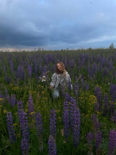 A young woman with wavy brown hair is kneeling in a field of tall purple lupine flowers and smaller yellow wildflowers. She is wearing a light-colored bomber jacket and blue jeans. She is holding a small bouquet of white daisies in her left hand. The sky above is overcast with dark, dramatic clouds, suggesting impending rain. The scene is set in a rural area, specifically identified as Kuyeda, Russia.