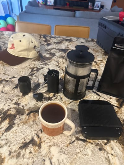 The image displays a collection of coffee-making equipment and related items arranged on a speckled granite or quartz countertop. No people are visible.

In the center, a clear glass French press with a black frame and plunger contains some residual liquid and grounds, suggesting it has been recently used. To its right, a black digital coffee scale is visible, featuring a textured surface and control buttons labeled "M", an up arrow, a down arrow, and "O/T" (likely On/Tare). A ceramic mug, light-colored with a patterned, light brown base, sits near the scale, filled with dark liquid, presumably coffee.

Below the French press, a black manual coffee grinder is shown disassembled, with its main body, handle, and a separate catch cup lying on the counter. To the left, a light-colored baseball cap with dark eyelets is placed, featuring an embroidered emblem and some indistinct red text, possibly a team name.

The background, largely out of focus, reveals an indoor setting, likely a living or dining area. Elements include a grey sofa, light brown upholstered chairs, and what appear to be colorful balloons, suggesting a casual, domestic environment. The scene implies a moment either during or after coffee preparation in a home in Mérida, Mexico.