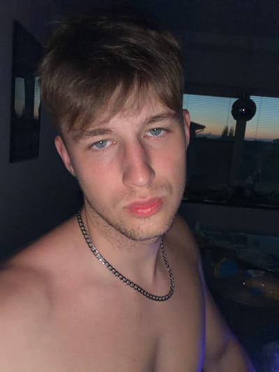 A young man with light brown hair and blue eyes is pictured in a close-up, chest-up selfie. He is wearing a silver chain around his neck. The background appears to be an indoor setting with a window showing a twilight sky and blinds. The lighting is dim, with some blue light reflecting on his chest. The man's expression is neutral, and he is looking directly at the camera. The image was taken indoors, likely in a residential setting.