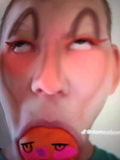 The media file is a close-up, low-resolution, and slightly blurry portrait of a person. The individual is facing forward and looking upwards, with their eyes rolled back. They have prominent makeup on, including bright orange eyeliner extending outwards from the eyes and arched dark brown eyeshadow or drawn-on eyebrows. There is also significant red blush applied to the cheeks. The person's mouth is open, revealing what appears to be a round, orange object inside. This object is decorated with drawn-on cartoonish eyes and rosy cheeks in pink. A faint watermark with text is visible on the right side of the image. The lighting appears to be artificial, casting a reddish hue over the subject's face. No specific setting or time of day is discernible. The overall impression is of a humorous or exaggerated self-portrait, possibly for social media. The text on the watermark appears to be a username or handle.