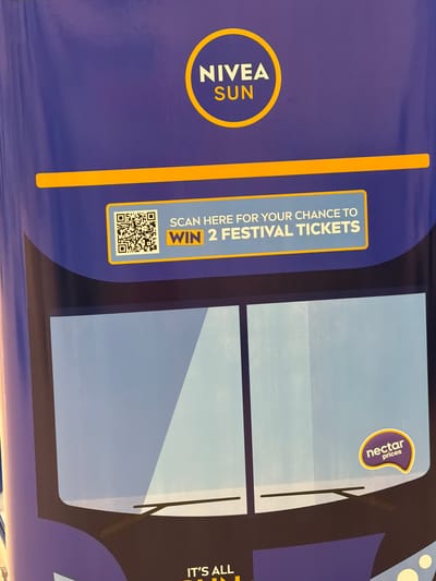 The image shows a portion of a bus advertisement in Pity Me, United Kingdom. The advertisement features the NIVEA SUN logo prominently on the left side. To the right of the logo, there is a call to action: "SCAN HERE FOR YOUR CHANCE TO WIN 2 FESTIVAL TICKETS," accompanied by a QR code for scanning. The central section of the advertisement displays an image that resembles the front windows and windshield wipers of a bus. In the upper right corner, there is a logo that reads "nectar prices". The right edge of the advertisement shows a partial text that reads "IT'S ALL". The background color is primarily a deep blue/purple. The advertisement seems to be promoting a contest or giveaway related to festival tickets in conjunction with NIVEA SUN and nectar prices.