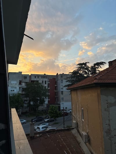 The image depicts an urban scene at sunset in Kyustendil, Bulgaria. The foreground is dominated by a concrete balcony railing, which casts a shadow across the lower portion of the frame. Beyond the railing, a street is visible with several parked cars, including a white Volkswagen Golf, a silver sedan, and a darker SUV. The street runs alongside a row of apartment buildings. The building directly behind the street is a multi-story structure with a red section on its right side and numerous windows. Another building, partially visible to the right, has a reddish-brown tiled roof and a weathered yellow facade with visible air conditioning units. In the background, a large evergreen tree with dark foliage stands against a sky filled with dramatic clouds illuminated by the setting sun, creating a warm, golden hue. The overall impression is one of a residential urban environment during twilight.
