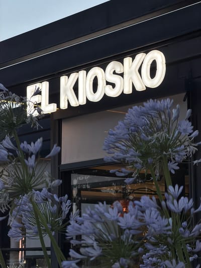 The image shows a close-up of illuminated white letters spelling "EL KIOSKO" on a dark building facade. In the foreground, a cluster of pale blue flowers with thin petals and long stems is in focus, obscuring the lower part of the sign and a window behind it. The building appears to be a commercial establishment, possibly a cafe or shop, given the signage. The setting is outdoors, and the lighting suggests it is either dusk or early morning. The overall impression is one of a modern establishment with natural elements. The location of Majadahonda, Spain is mentioned but not visually verifiable from the image content.
