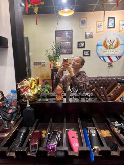 The image shows a woman taking a selfie in a barber shop. She is holding a mobile phone and appears to be looking at its screen. She is wearing a hooded sweatshirt. In the background, there is a mirror reflecting the shop's interior, which includes a barber pole, framed pictures on the wall, and various barber tools such as clippers and brushes. There are also decorative items like a golden cat statue and figurines on a shelf. The shop appears to be located in Xuân Hương - Đà Lạt, Vietnam, as indicated by the Vietnamese text on a sign in the mirror and the style of the decor. The lighting suggests it is daytime.