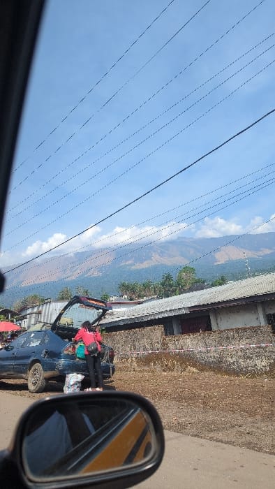 This image depicts a roadside scene in Buea, Cameroon, with Mount Cameroon visible in the background under a blue sky with scattered clouds.
In the immediate foreground, the black side-view mirror of a vehicle is visible, reflecting a yellow and black striped object and a partial white object.
In the midground, a dark blue sedan is parked on the right side of a dirt road. The vehicle appears dirty, with the rear left fender showing signs of damage or missing a fuel cap. Its trunk is open. A female individual, dressed in a red long-sleeved top, dark pants, and light-colored shoes, is leaning into the open trunk, appearing to handle items. She wears a dark crossbody bag. A large, multi-colored patterned bag rests on the ground directly behind the sedan.
Adjacent to the dirt road, on the right, is a low, rough concrete or stone wall with a segment of red and white striped tape affixed to it. Behind this wall is a single-story building with a gray corrugated metal roof and discolored walls. To the left, a segment of a red umbrella or awning is visible.
The background features Mount Cameroon, with its lower slopes covered in dense green forest, transitioning to large patches of brown, sparsely vegetated terrain at higher elevations. The mountain's peak is partially obscured by white clouds. Numerous overhead power lines extend across the blue sky. Scattered structures and tropical foliage are visible on the lower mountain slopes.