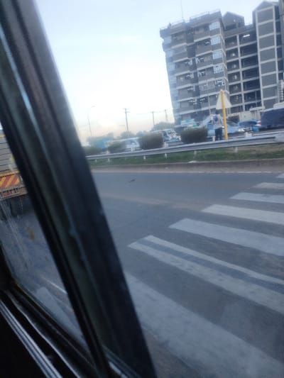 The image displays an urban road scene in Murera ward, Kenya, observed from a vehicle interior, with a window frame visible on the left and bottom. A dark asphalt road with a white-striped pedestrian crossing occupies the lower portion of the frame. On the far left, the rear section of a truck with red and yellow reflective stripes is partially visible.

In the midground, a multi-lane roadway features a central median strip containing small shrubs and a metal guardrail. Multiple vehicles, including cars and a light-colored minibus (matatu), are present on the road. One individual, dressed in a light-colored top and dark trousers, stands on the median strip next to a yellow triangular road sign. Overhead utility poles and streetlights are also visible.

The background is dominated by a multi-story building on the right side, characterized by a grey facade and numerous windows, suggesting commercial or residential use. The sky is light and clear.