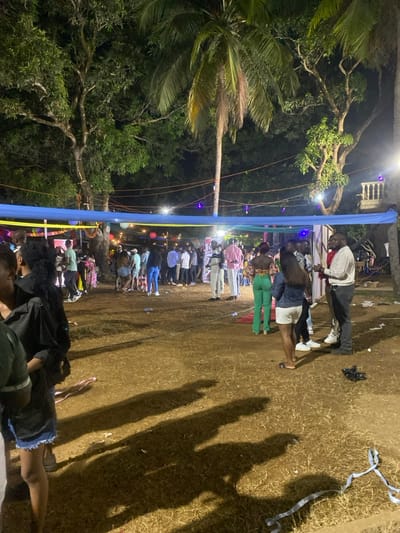 This media file captures an outdoor social gathering at night in Freetown, Sierra Leone. The scene is brightly lit by artificial lights, including spotlights and string lights, which create long, distinct shadows of the attendees on the earthy ground.

Numerous people, mostly young adults, are present. They are seen standing, mingling, and observing, suggesting a relaxed and communal atmosphere. Some individuals in the foreground appear to be sitting or lounging.

A prominent blue fabric banner, possibly with yellow accents, stretches horizontally across the middle of the frame, serving as a decorative element or part of a temporary structure. String lights with small, glowing bulbs are visible, strung along this banner and between the surrounding trees. Large tropical trees, including tall palm trees, flank the gathering space, indicating a natural, open-air setting.

The ground is dry, possibly dirt or sparse grass, with some scattered debris resembling streamers or confetti. In the background, a white drum is faintly visible, hinting at music or entertainment. There is no visible text in the image. The overall impression is that of an evening event or celebration under the open sky.