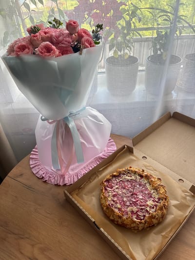 A bouquet of pink roses and carnations sits on a wooden table next to an open cardboard box containing a rustic galette topped with berries and sliced almonds. Behind the table, a window overlooks a city, with potted plants visible on the windowsill and sheer white curtains softening the light. The scene appears to be set indoors, possibly in a home or shop, on a bright day. The style of the bouquet and galette suggests a gift or a celebratory treat.