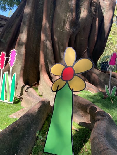 The image depicts a decorative display of cartoon-like flowers placed in front of a massive tree trunk with gnarled roots. The flowers are flat, brightly colored cutouts. The largest flower, positioned centrally, is yellow with a red center and a green stem. To the left, two pink, spiky flowers stand with green stems. To the right, a single pink, spiky flower with a green stem is visible. The background is dominated by the dark, textured bark of a very large tree. The ground is covered in green artificial turf, with fallen leaves and twigs scattered on it. Sunlight casts shadows on the tree and the ground. This scene appears to be an outdoor decoration, likely in a public space or park. Based on the information provided, the location is the city of Ciwaruga, Indonesia. There are no people or animals visible in the image.