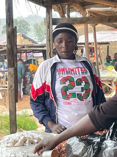 This image depicts a busy marketplace scene in Wang, Nigeria. The primary subject is a person wearing a white t-shirt with "UNLIMITED" printed vertically on the left side and a large graphic of the number "33" in red and green. The t-shirt is layered under a jacket with red and white stripes on the sleeves. The person appears to be looking down with a neutral expression.
Behind the main subject, other people are visible in the background, some tending to stalls or interacting with customers. The setting is rustic, with wooden structures providing shelter and support for the market. Bags of produce, possibly peanuts, are visible in the foreground on the right side of the image, contained within a clear plastic bag and a dark plastic bag. A small tin can is also present near the produce. The overall atmosphere suggests a typical day at a local market, possibly in the morning or afternoon, with the weather appearing overcast. The presence of trees and a hilly landscape in the distance provides a glimpse of the natural surroundings of Wang, Nigeria.
Visible text includes "UNLIMITED" and "YOUR YOUNG GENERATION".