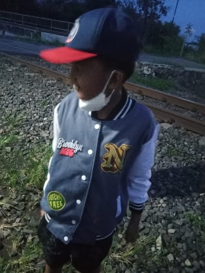 The image captures a young individual standing near railway tracks in Sidorejo, Indonesia, likely during dusk or early night due to the low light. The person is seen from the chest up, looking to their left. They are wearing a dark blue baseball cap with a red brim, and a white face mask covers their nose and mouth.
The main subject is dressed in a grey or light blue varsity-style jacket with white sleeves and white striped cuffs. The jacket has several patches: on the left chest, "Brooklyn NYC" is visible, with "Brooklyn" in white script and "NYC" in red below it. On the right chest, there is a large, stylized letter "N" in yellow/gold with a dark outline. Near the bottom left of the jacket, a circular patch reads "FOREVER FREE" in yellow text within a green background. White buttons run down the front of the jacket. Dark shorts are partially visible below the jacket.
The ground around the person is covered in railway ballast (crushed stone) and patches of grass or weeds. The railway tracks extend into the distance. In the background, a road, a bridge or overpass with railings, and dense dark foliage or trees are visible under a dark sky. A distinct white X-shaped sign, characteristic of a railroad crossing, can be seen in the background on the right side. The overall scene suggests a casual setting near a railway line.