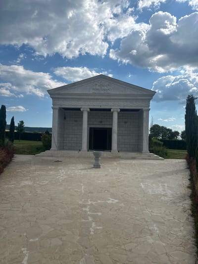 The image depicts a grand, white marble building with a classical architectural style, resembling a temple or mausoleum. It features a prominent pediment adorned with relief sculptures, a row of Ionic columns supporting a portico, and walls constructed of large, rectangular stone blocks. A wide, paved pathway made of irregularly shaped flagstones leads directly to the building's entrance, which is a dark, open doorway. In the center of the pathway, in front of the entrance, stands a small, decorative stone urn or pedestal.

The building is situated within a meticulously landscaped area. Tall, slender cypress trees line the sides of the pathway, framing the view of the structure. Beyond the building, there are rolling green hills and trees under a bright blue sky filled with dramatic, puffy white clouds. The overall impression is one of solemnity and grandeur, set against a natural, serene backdrop. The location is identified as the city of Neofit Rilski, Bulgaria. There are no people or animals visible in the image.