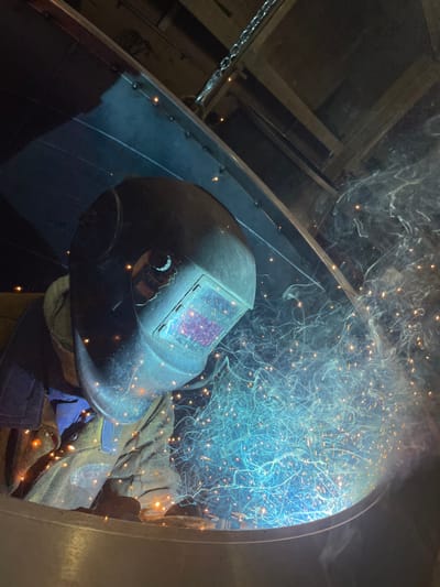 A person wearing a welding helmet and protective clothing is engaged in welding, creating a shower of sparks and smoke. The welding appears to be taking place inside a large, cylindrical metal structure. The location of Tomba, Montenegro, is not discernible from the image.