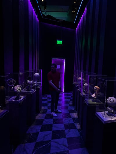 A man stands in a dimly lit hallway adorned with vertical black and purple striped walls. He is wearing a red t-shirt, dark pants, and white sneakers. The hallway features several glass display cases on either side, showcasing various sculpted figures and props, some of which appear to be character heads and puppets. The floor is a black and white checkered marble pattern. An illuminated green "exit" sign is visible on the back wall. The scene appears to be an exhibition or museum display, possibly related to animation or film, and based on visual cues of the exit sign and architectural style, it is highly likely to be located in the Cintermex convention center in Monterrey, Mexico, specifically within a Tim Burton-themed exhibition.