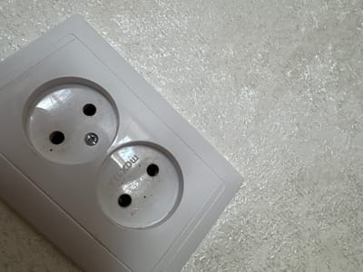 A light-colored, double electrical socket is mounted on a textured, speckled wall. The socket is oriented diagonally within the frame, with the top left corner of the socket plate closer to the camera than the bottom right. The wall surface appears to be wallpaper with a subtle, reflective pattern, possibly metallic or glittery. One of the socket openings has a small screw visible, and text that reads "max 16A" is visible on the inner rim of the other socket opening. There are no people or animals present in the image. The scene is indoors, likely in a residential or commercial building. There is no context provided to determine the specific location within the city of Shchuchinsk, Kazakhstan.