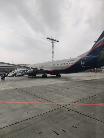 The image depicts an Aeroflot Boeing 737-800 aircraft, registration RA-73189, parked on the tarmac at Yemelyanovo Airport (KJA) in Krasnoyarsk Krai, Russia. The aircraft is a passenger jet with its livery featuring the Aeroflot logo and the Russian flag colors on the tail. A mobile airstair is connected to the aircraft's forward cabin door, and several passengers are visible disembarking or boarding the plane. In the background, airport buildings and a control tower or lighting mast are present under a cloudy sky. The tarmac is made of concrete with painted red and black lines. Two individuals are standing on the tarmac in the foreground, observing the aircraft.