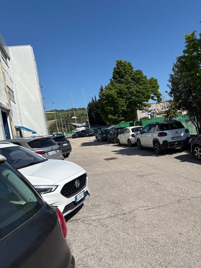 This image depicts an outdoor parking lot under a bright, clear blue sky, characteristic of a sunny day. The photograph is oriented vertically (rotated 90 degrees clockwise).

Numerous cars are parked in rows on an asphalt surface, which shows some texture from the sun's glare. In the foreground on the right, a white Nissan Qashqai (or similar SUV) is visible, facing left, with its Italian license plate "DJ 847 FF" clearly readable. Adjacent to it, the rear of a dark grey car with a prominent red taillight is partially seen. Further along the row, another grey car displays the Italian license plate "AN 099 HGT," where "AN" signifies Ancona, Italy. Towards the center, a white Citroën C3 Aircross is parked, with its "Citroën" logo and Italian license plate "GV 277 GR" visible on the rear. Several other vehicles, mostly dark, are parked further down the rows.

On the left side of the image, a plain white building with several rectangular windows and some exterior piping is present. Green trees with dense foliage are scattered throughout the scene, casting sharp shadows on the ground, indicating the strong sunlight of daytime, likely mid-day or early afternoon. In the background, beyond the cars and trees, a distant landscape of hills or fields is visible under the expansive blue sky. Several tall, slender white poles are also discernible in the middle distance.

No people or specific activities are observable in the image, which primarily focuses on the parked cars and their surroundings in Ancona, Italy.