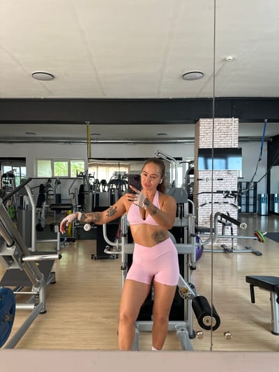 A woman with tattoos on her arm, wearing a pink sports bra and pink shorts, is taking a mirror selfie in a gym. She is holding a phone in her right hand and wearing white gloves. The gym is equipped with various weight machines, a bench, and weights. The floor is made of light-colored wood, and the walls are white with some brick accents. The ceiling has circular lights and exposed pipes. The image is a reflection in a large mirror.