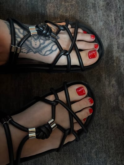 The image shows a view of a person's feet clad in black gladiator-style sandals with red painted toenails. The sandals feature a crisscrossing strap design with decorative knots and silver metallic accents. The right foot exhibits a tattoo on the instep, primarily in black ink. The background consists of a dark, textured surface, possibly a rug or carpet. The location is potentially in West Hollywood, United States of America, though there are no readily identifiable landmarks within the frame. No specific actions or interactions are visible.