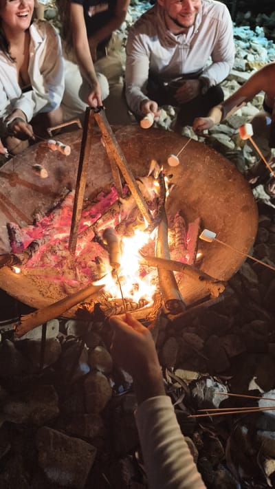 A group of people are gathered around a bonfire, roasting marshmallows. The scene appears to be outdoors, possibly a campsite or a backyard in Cam Lam, Vietnam. The fire is burning brightly in a large metal basin, with logs and embers visible. Several individuals are holding sticks with marshmallows over the flames, with some marshmallows already toasted to a golden brown. The people are casually dressed, and some are smiling and laughing, indicating a relaxed and enjoyable atmosphere. The ground around the fire is covered in rocks and dry leaves.