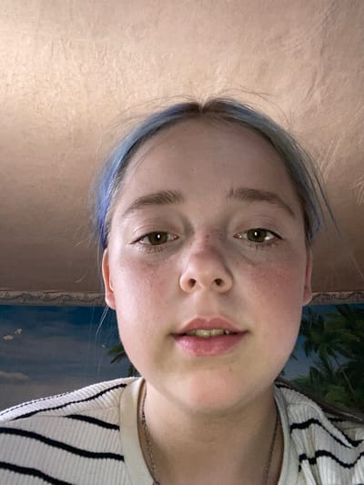 The image is a close-up, frontal, selfie-style photograph of a young female. She has fair skin with freckles, light brown eyes, and light brown hair with streaks of blue. She is wearing a white and black striped shirt and a silver necklace. The background appears to be a wall with a textured finish and a mural of a tropical scene with palm trees and a sky with clouds. The lighting is soft and natural. The image is sharp and clear.
There is no information in the image to suggest a specific location in the city of Nova Praha, Ukraine.