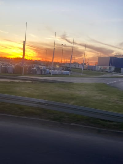The image depicts an outdoor industrial or commercial area at sunset in Bielany Wrocławskie, Poland. The sky is ablaze with vibrant orange and yellow hues, indicating a dramatic sunset. Tall, slender poles, likely streetlights or flagpoles, are prominent in the foreground and midground, some with illuminated tops. In the midground, there is a large parking area or lot filled with stacked materials, possibly construction supplies like bricks or paving stones, neatly arranged in rows. Several vehicles, including what appears to be a truck with the word "Raben" visible on its side, are parked in the lot or driving on a road. Behind the stacks of materials, a large, modern building with blue and gray elements stands, suggesting a warehouse, distribution center, or industrial facility. The scene is bordered by a grassy area and a road with a guardrail, implying a thoroughfare or access road adjacent to the industrial site. The overall impression is one of commercial activity set against a striking natural sky phenomenon.