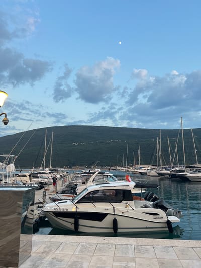 The image depicts a marina in Đenovići, Montenegro, likely in the late afternoon or early evening given the soft light and the presence of the moon in the sky. The sky is a light blue, with scattered white and gray clouds. A lush green, tree-covered mountain range dominates the background, rising steeply from the coastline.
In the foreground, several boats are docked along a stone pier. The closest boat is a white motorboat with a black trim and two outboard motors. It has the registration "POL001SFM" clearly visible on its side, and the brand "QUICKSILVER" is also legible. Another boat, also white, is visible behind it, with the name "CLARA 3" written on its side, and a small red and white flag attached. Further down the marina, numerous sailboats with tall masts are moored, creating a dense arrangement of rigging against the mountainous backdrop.
There are a few people visible on and around the boats, though their features are not distinct. One person in a red shirt appears to be standing on a boat in the middle ground. The water in the marina is calm and reflects the surrounding boats and sky. The overall scene suggests a peaceful atmosphere at a popular boating destination.