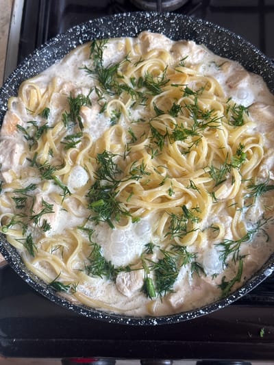 A close-up overhead shot shows a creamy pasta dish being prepared in a black speckled pan. The dish appears to be fettuccine or linguine tossed in a rich, white sauce with chunks of chicken visible throughout. Fresh dill is sprinkled liberally over the pasta, adding a vibrant green contrast. Bubbles rise from the sauce in several places, indicating that the dish is actively cooking. The pan is set on a black stovetop, with a hint of a gas burner visible at the bottom left. The lighting suggests an indoor setting, likely a kitchen. The focus is solely on the food, with no people or other elements of the surroundings clearly visible. The scene is one of home cooking, with the preparation of a delicious meal in progress. The time of day, weather, and specific location cues beyond being in a kitchen are not discernible. No text is visible in the image.