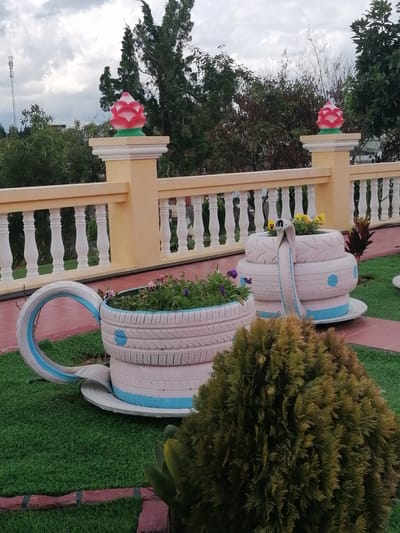 In the city of Lộc Quý, Vietnam, an outdoor scene features two large planters made from painted tires, designed to resemble teacups. The teacup planters are filled with green plants and some purple and yellow flowers. One planter, larger than the other, is made from three stacked tires and has a spout-like feature made from a tire segment. It rests on a saucer-like base, also made from a tire. The smaller planter, shaped like a teacup with a large handle made from a tire segment, contains green plants. Both planters are painted white with light blue accents, including circular patterns on the stacked planter and a stripe on the handle of the teacup planter.
In the background, a beige, ornate railing with white balusters runs along a raised terrace. Beyond the railing, trees and foliage are visible under a cloudy sky. Two decorative lotus-shaped finials, one red and white with a green base, sit atop pillars supporting the railing. To the far left, a tall telecommunications tower is visible against the sky. The ground in the foreground is covered with artificial green turf, bordered by red brick. A dense green shrub is positioned in the lower right corner of the image.